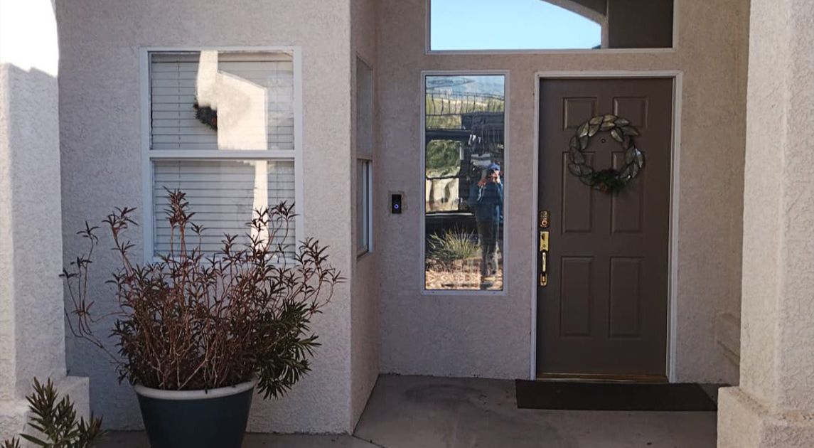 Front porch of a residential home featuring a security screen door with a wreath, flanked by stucco walls and a large potted plant.